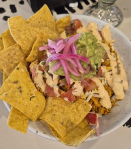 A bowl with shredded chicken, cheese, tomatoes, creamy sauce, guacamole, and pickled onions.