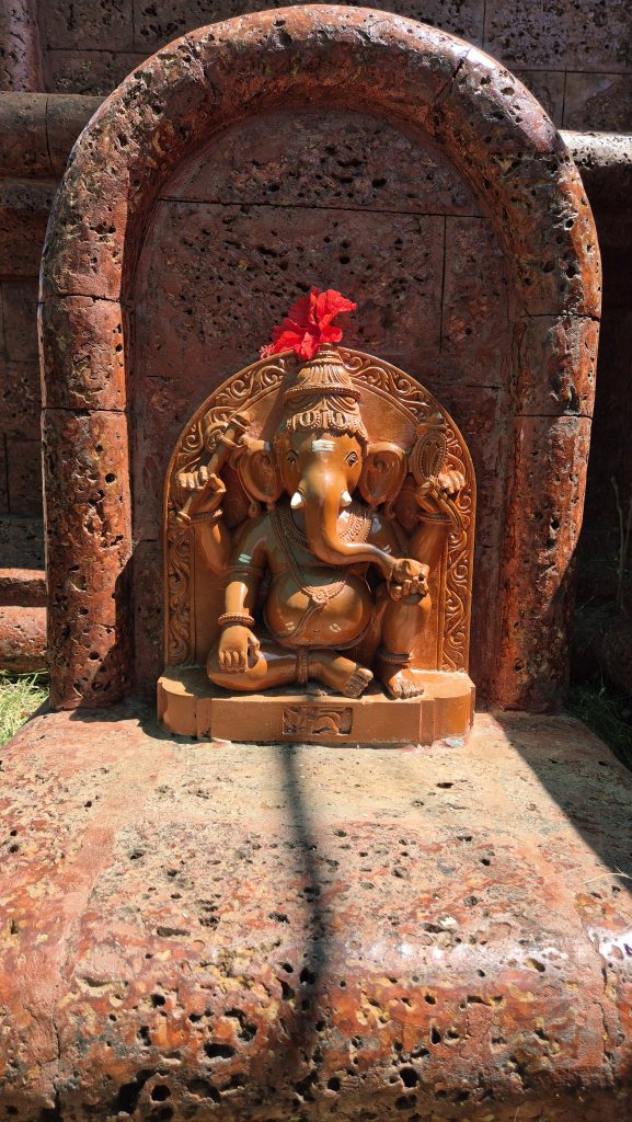 A small reddish-brown statue of Ganesha, the elephant-headed god with four arms holding symbols, sits on a stone base with a red flower on his head, against old textured stone in soft sunlight.