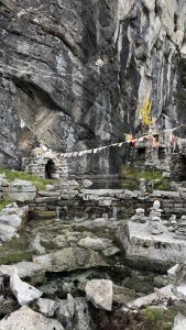 A rocky cliffside features a small stone shrine adorned with colorful prayer flags. 