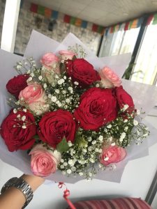 Hand holding a bouquet of red and pink roses with white baby's breath, wrapped in white paper, against a softly blurred colorful indoor background