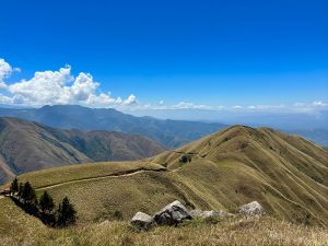Fotografía de una serie de montañas cubiertas de pasto. En primer plano se observan rocas y vegetación seca. Al fondo se distinguen más montañas y un cielo azul con nubes blancas en el lado izquierdo.