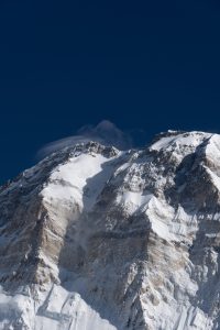 Steep rocky mountain ridge with patches of snow and ice against a dark blue sky, with wind-blown snow trailing off the summit from Nepal