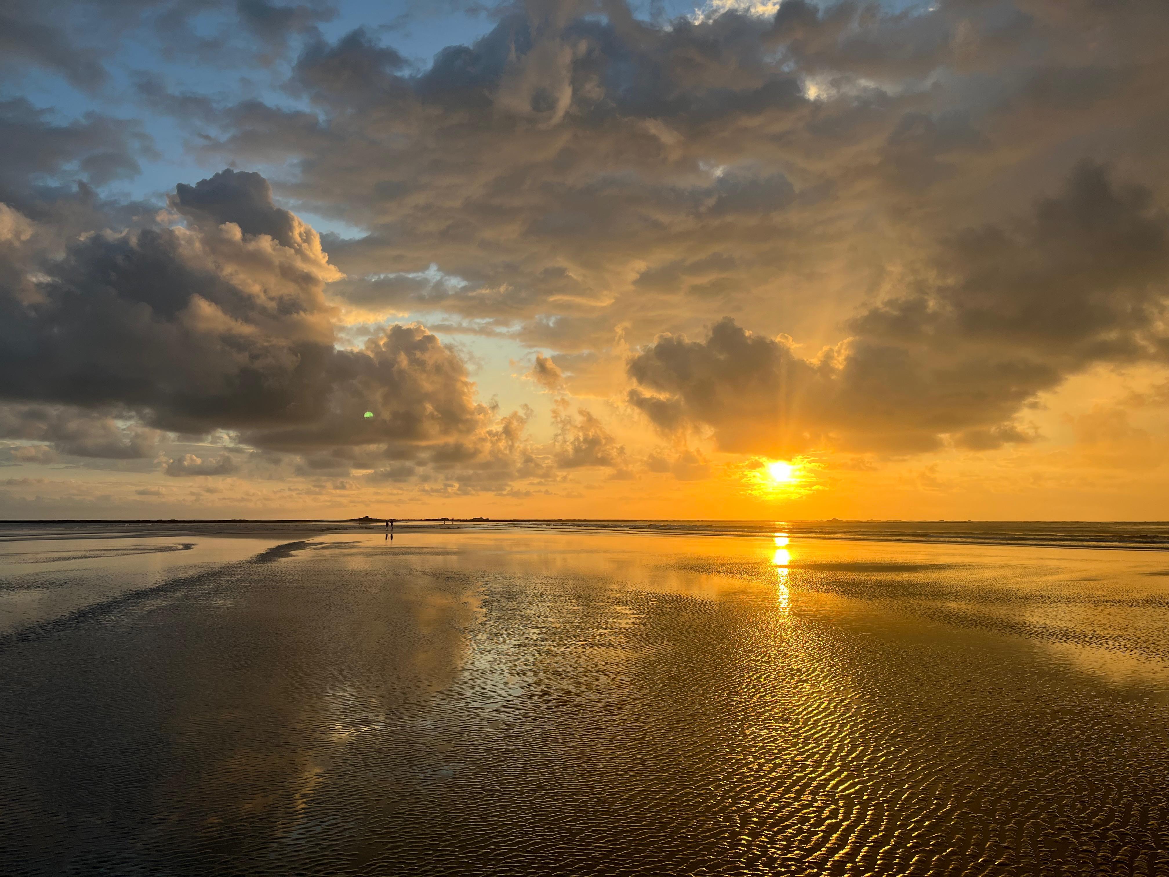 Sunset beach with reflective wet sand, colorful clouds, and two people walking along the shore.