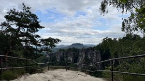 A scenic mountain viewpoint with a metal railing overlooking dramatic vertical rock pillars rising from a dense forest, framed by pine and leafy trees beneath a cloudy sky.
