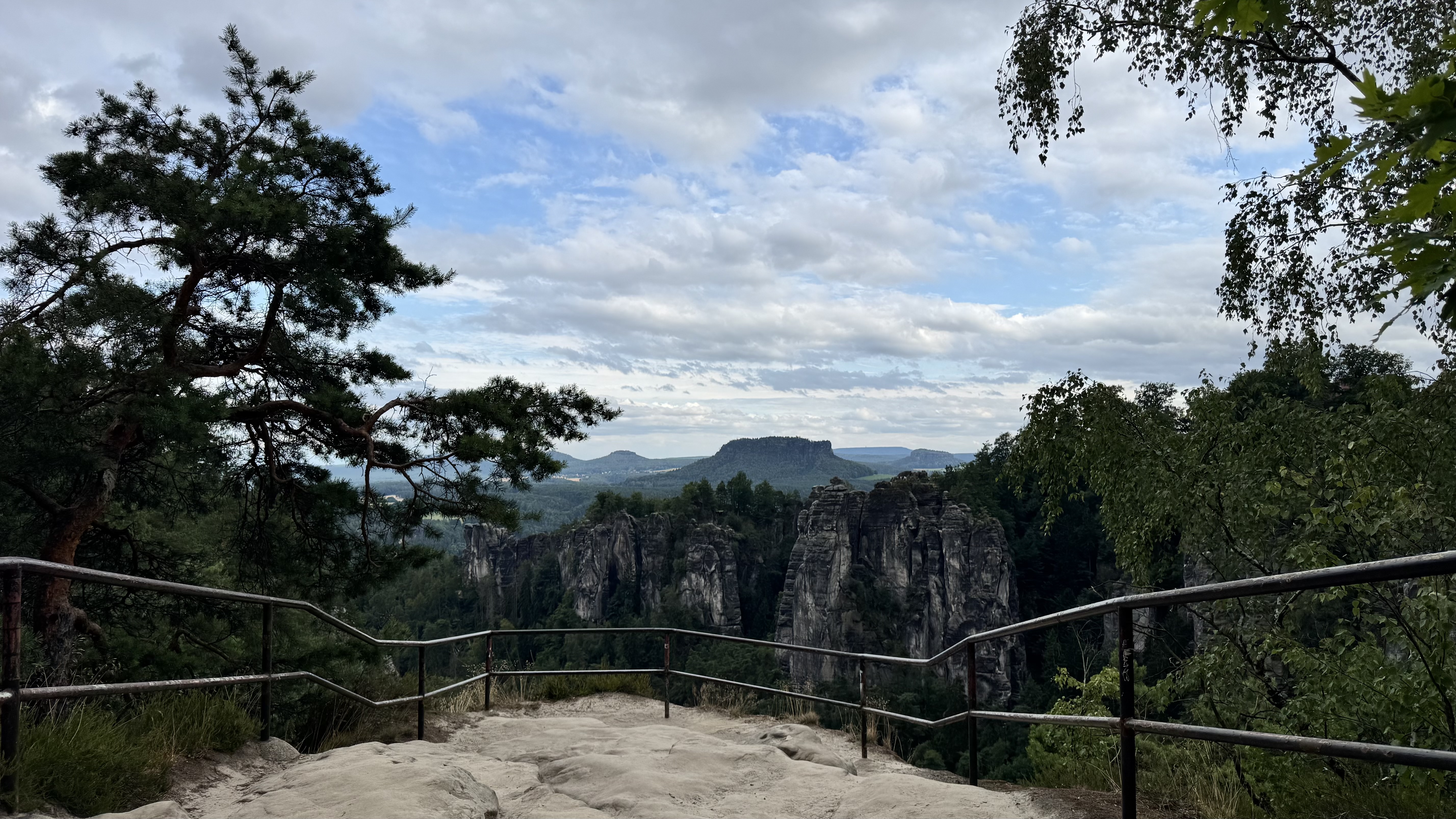 A scenic mountain viewpoint with a metal railing overlooking dramatic vertical rock pillars rising from a dense forest, framed by pine and leafy trees beneath a cloudy sky.