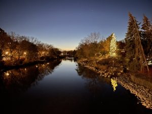 A calm river at dusk reflects dark trees, glowing park lights, and a brightly illuminated evergreen on the right bank. Bare branches and tall evergreens frame the water, while a deep blue evening sky with a few visible stars stretches overhead. Rocky shoreline and warm reflections create a peaceful night scene.