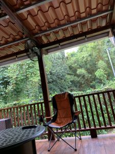 A covered wooden porch with a terracotta-tiled roof, a folding orange and gray camping chair, a wall-mounted fan, and a metal table, overlooking a dense bamboo and green forest through a wooden railing.