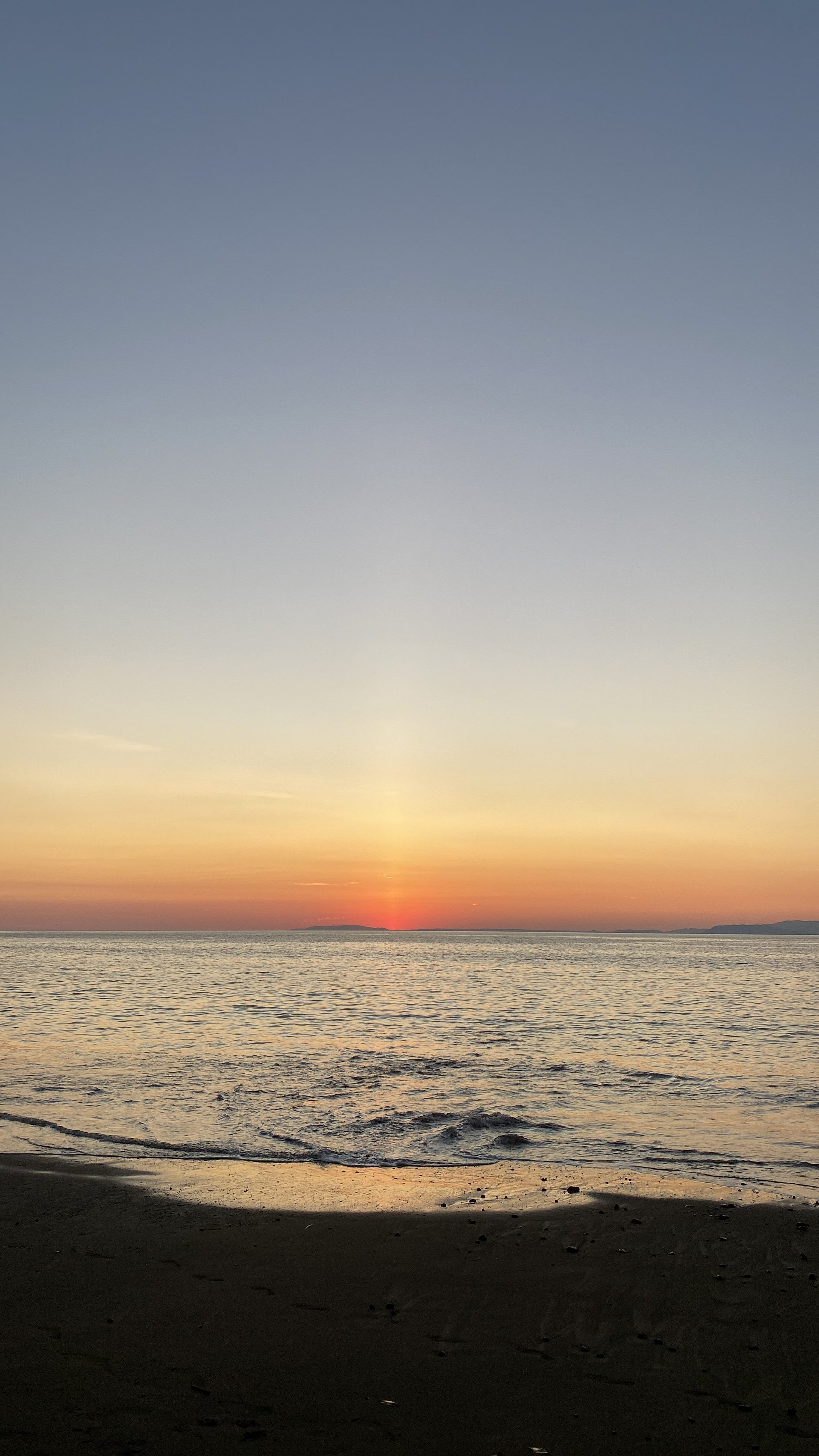 A tranquil beach scene at sunset, with gentle waves lapping at the shore. The horizon displays a gradient of colors, transitioning from soft blues in the sky above to warm oranges and reds nearer to the water, where the sun is just dipping below the horizon. The calm surface of the ocean reflects these hues, creating a serene and peaceful atmospher