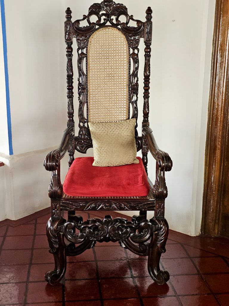 An antique wooden chair with detailed carvings, a woven cane backrest, and a bright red cushion reflects the elegance of traditional craftsmanship. Captured at Mangode, Puthunagaram, Palakkad.
