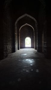 A dark stone corridor leads towards a bright opening at the far end, framed by an archway. 
