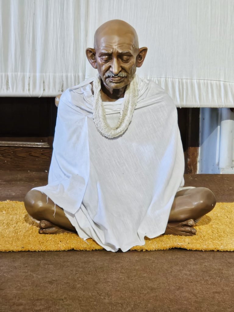 A peaceful statue of Mahatma Gandhi sits in a meditative pose, dressed in his simple white shawl, reflecting his values of truth, simplicity, and non-violence. Captured inside Aga Khan Palace, Pune, Maharashtra.