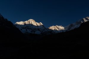 Snow-capped mountains at dawn cast dramatic shadows against a deep blue sky. Sunlight highlights the peaks, creating a serene and majestic atmosphere.