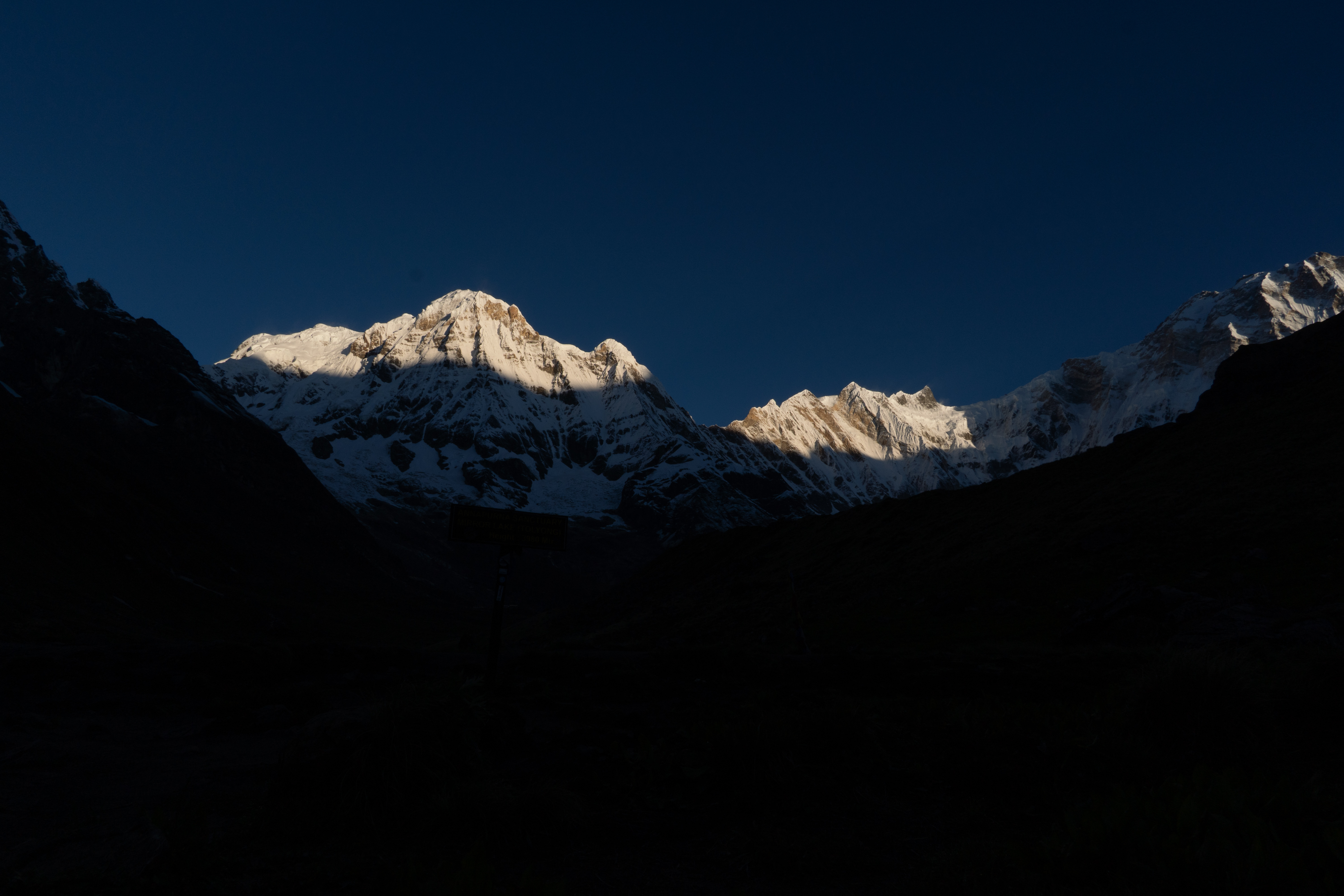 Snow-capped mountains at dawn cast dramatic shadows against a deep blue sky. Sunlight highlights the peaks, creating a serene and majestic atmosphere.