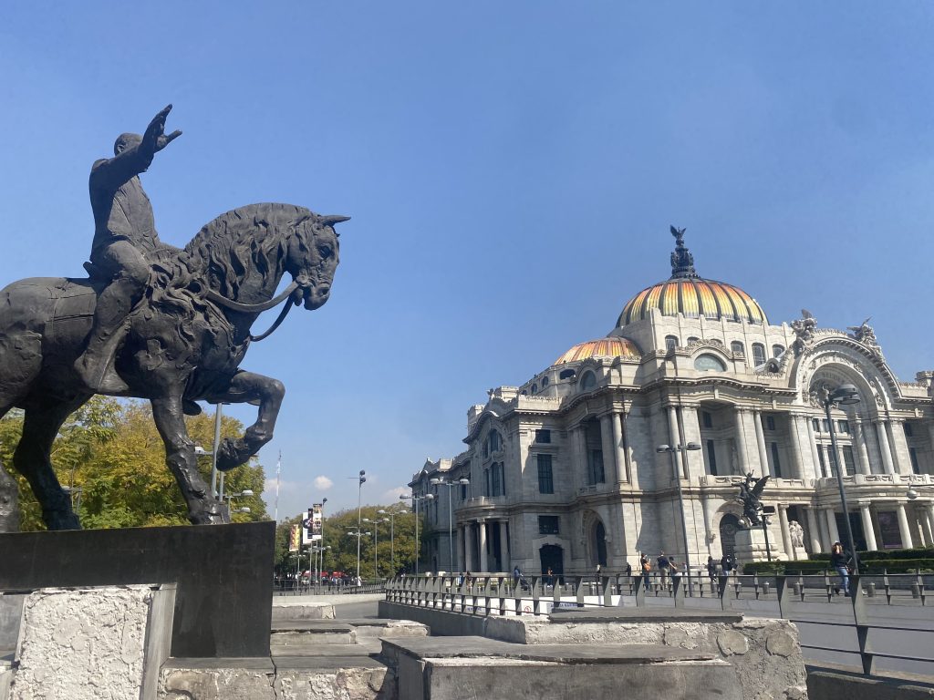 Palace of Fine Arts in Mexico City featuring its iconic colorful dome and marble facade, viewed alongside an equestrian bronze statue under a clear blue sky.