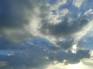 A wide-angle shot of a blue sky with heavy dark and white clouds, where the bright sun is partially hidden, creating a glowing silver lining and shining light from behind the clouds.
