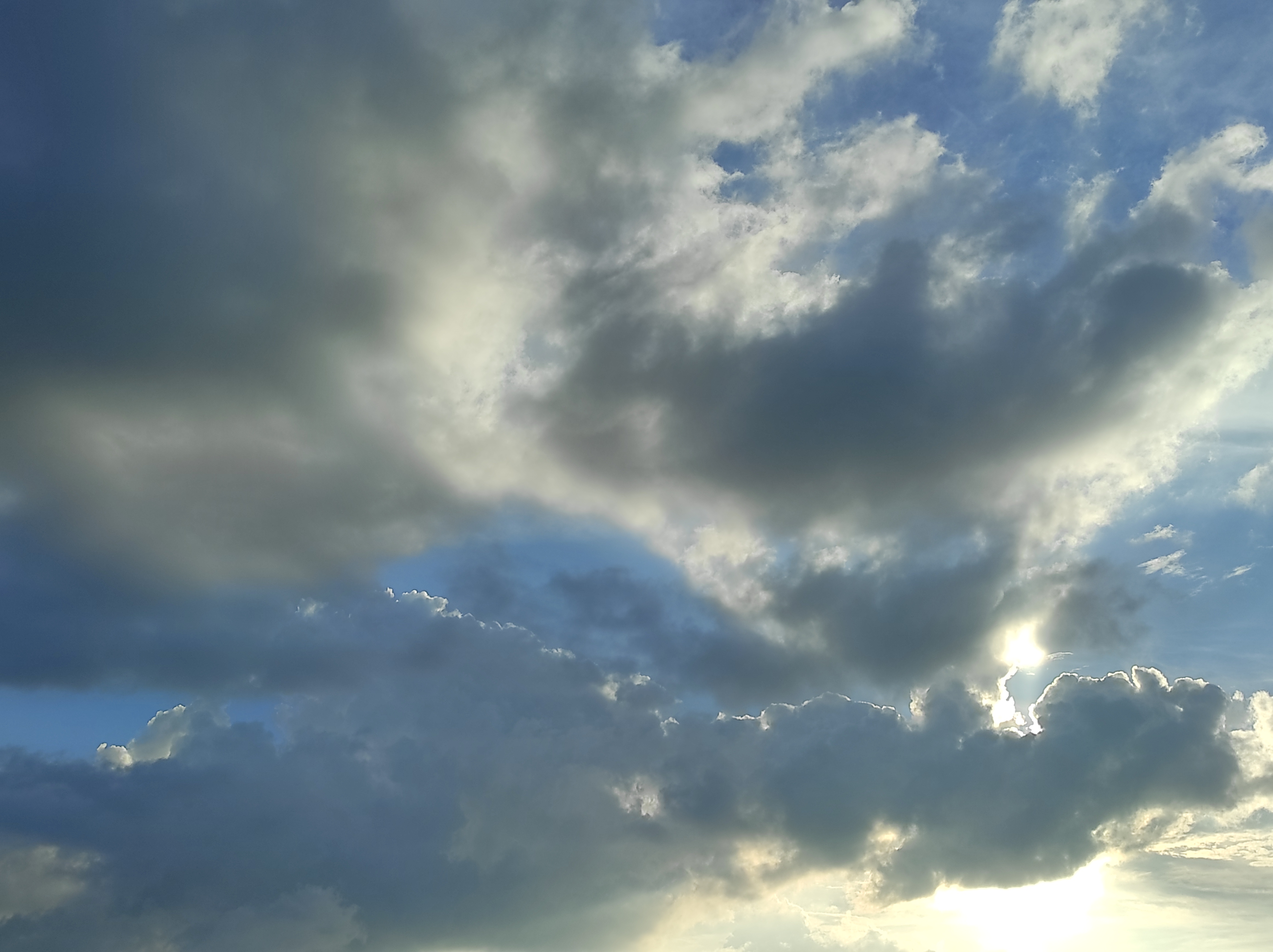 A wide-angle shot of a blue sky with heavy dark and white clouds, where the bright sun is partially hidden, creating a glowing silver lining and shining light from behind the clouds.