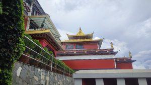 Colorful multi-tiered monastery with red walls, golden details, green bushes, and a stone path under a bright sky.
