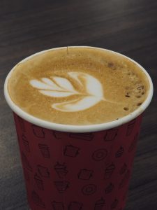 A freshly brewed latte is served in a paper cup, topped with elegant leaf-shaped latte art. The smooth coffee and creamy foam create a warm and inviting café moment. Captured in Palazhi, Kozhikode.