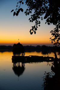 Silhouette of a small pavilion and wooden boat on a calm lake at sunrise, reflected in the water, with tree branches framing the scene.