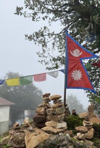 Nepal flag with stone cairns and colorful prayer flags in a misty setting.
