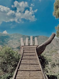 A large wooden hand sculpture with outstretched fingers forms a pathway against a backdrop of lush green mountains and a bright, cloudy blue sky.