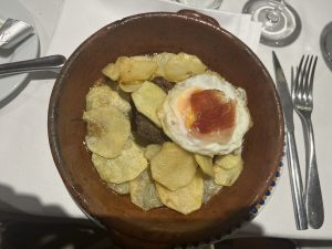 A round clay dish filled with a bed of golden, crispy potato chips arranged around a piece of beef. Atop the beef is a perfectly cooked fried egg with a runny yolk, and a small slice of cured ham resting on the egg. 