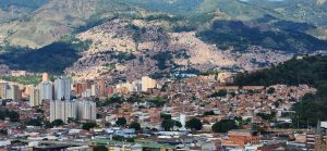A panoramic view of a vibrant urban landscape surrounded by mountains. In the foreground, there are various buildings of different heights and styles, with some high-rise apartments featuring a mix of white, yellow, and brick-colored facades