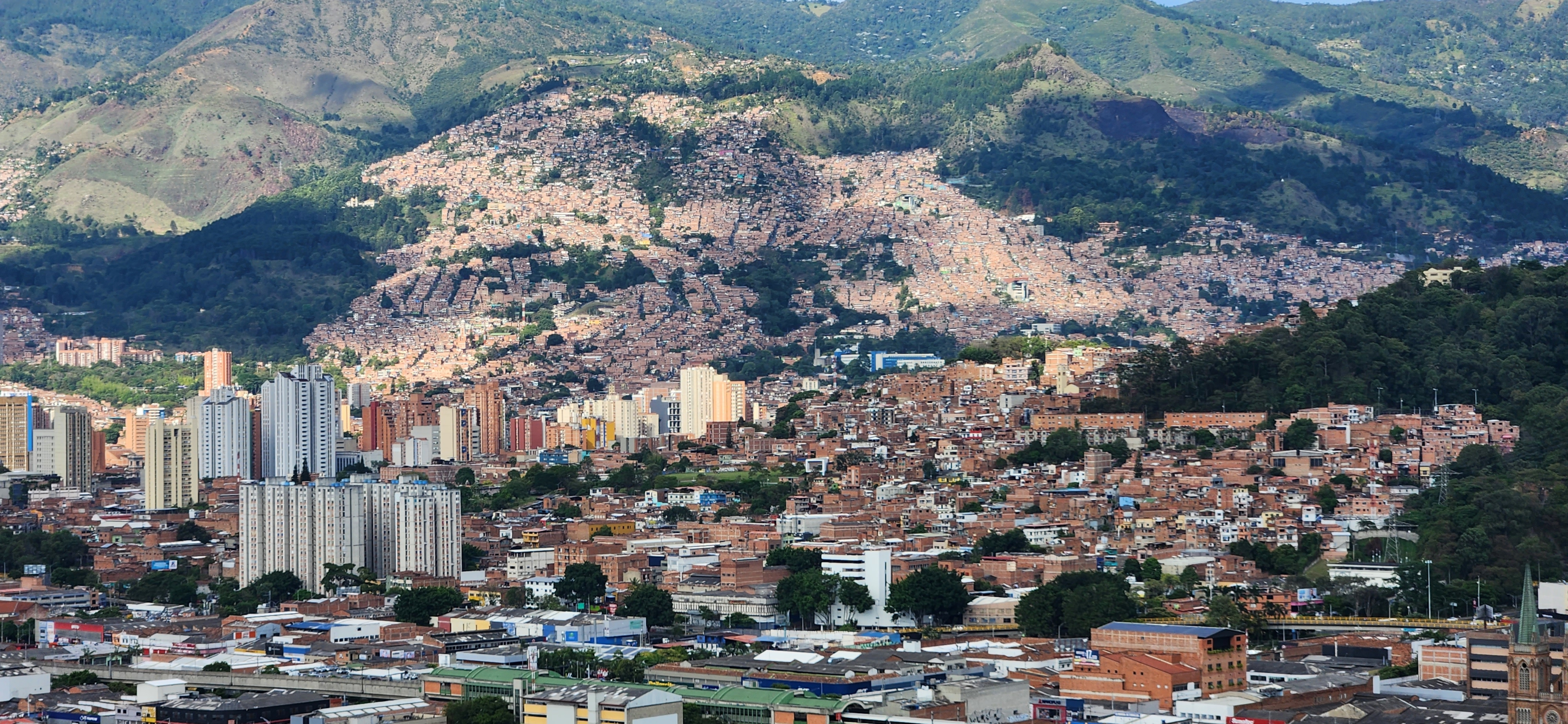 A panoramic view of a vibrant urban landscape surrounded by mountains. In the foreground, there are various buildings of different heights and styles, with some high-rise apartments featuring a mix of white, yellow, and brick-colored facades