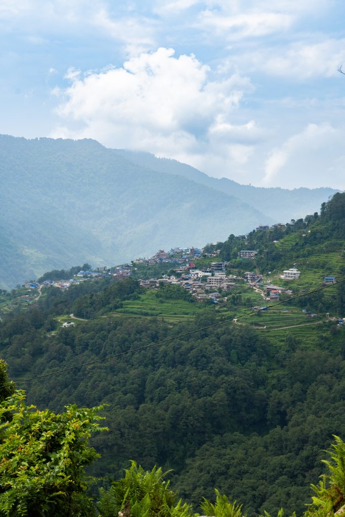 A lush, green mountain landscape with a small village on terraced hillsides, under a cloudy blue sky.