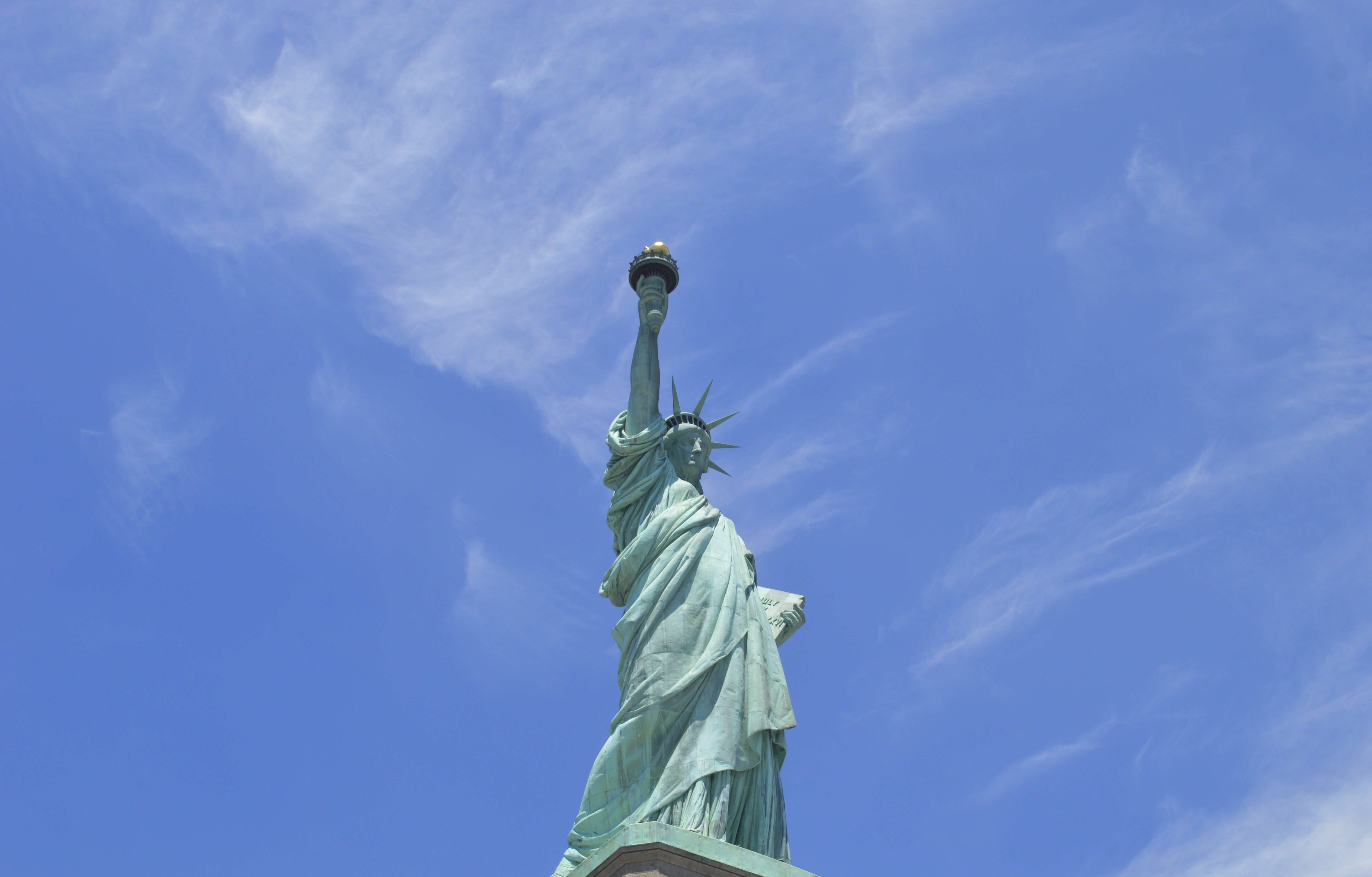 Low-angle view of the Statue of Liberty against a bright blue sky with thin, wispy clouds.