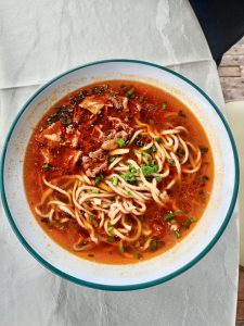 A bowl of Chinese noodles with soup topped with shredded meat, mushrooms, chopped spring onions, and broth.
