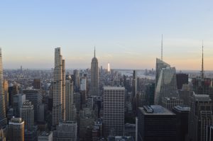 Elevated view of a dense city skyline filled with high-rise buildings.  A tall skyscraper stands near the center while the evening sun reflects off distant buildings beneath a pale blue sky