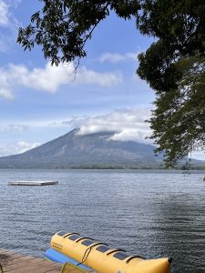 View of a calm lake with gently rippling water and a green volcano partially covered by white clouds in the background. A yellow kayak rests on a wooden dock in the foreground, while leafy branches frame the scene beneath a partly cloudy blue sky.