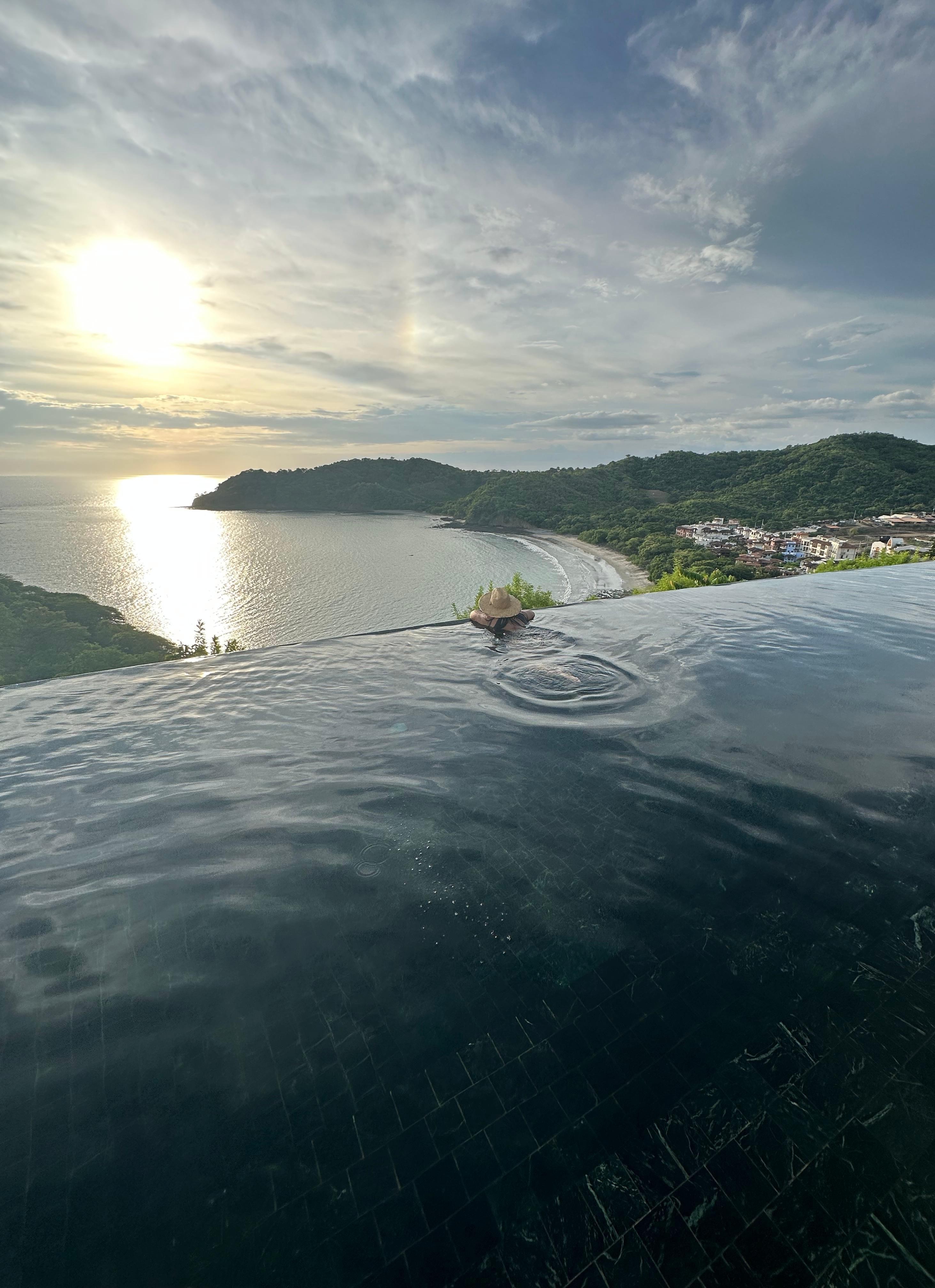 A serene view from a luxurious infinity pool overlooking a calm beach and rolling hills.