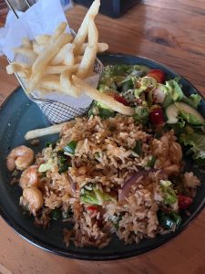 A dark green plate holds a serving of fried rice, which is mixed with various colorful vegetables such as broccoli, red bell peppers, and green onions. Juicy shrimp are scattered throughout the rice. On the side, there's a lightly dressed salad that includes leafy greens, slices of strawberries, and sesame seeds.