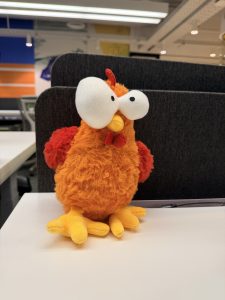 A plush orange chicken with exaggerated white eyes and a small red comb sits on a desk.
