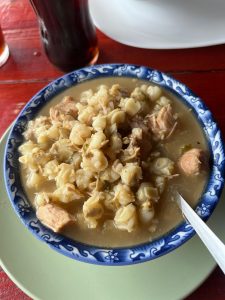 A bowl of hominy soup with meat in a light brown broth and a spoon on the side.