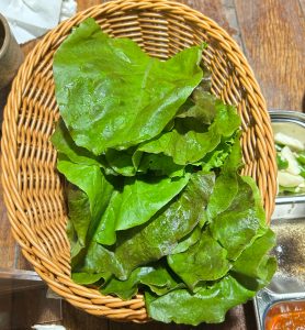 Fresh green lettuce leaves in a woven basket on a wooden table beside a small bowl and napkins.