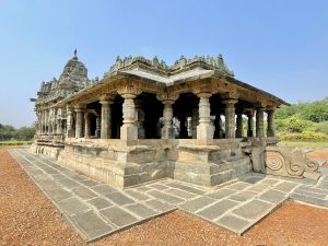 A beautifully preserved historic stone temple (Brahma Jinalaya) stands under a clear blue sky. The open-pillared hall, detailed carvings, stone pavement, and decorative base show the fine craftsmanship of Western Chalukya temple architecture. Captured in Lakkundi, Gadag district, Karnataka.
