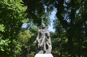 A bronze statue of a seated man holding a book and a pen on a stone pedestal. The monument is surrounded by tall leafy trees, with sunlight filtering through the dense green canopy
