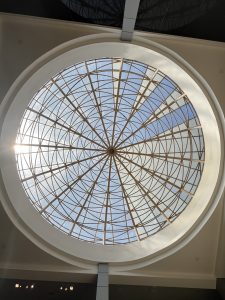 A large circular skylight fills the center of the photo, set within a white arched ceiling. Its intricate bronze-colored metal framework radiates from a central hub, forming concentric rings and geometric patterns against a blue sky. Sunlight streams through on the left.