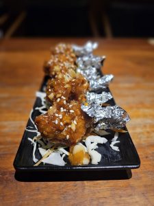 Fried chicken lollipops coated in a glossy, savory sauce and topped with sesame seeds. They are neatly arranged on a long black plate over shredded cabbage, with their bone handles wrapped in foil. Location: Edavannappara, Malappuram, Kerala.