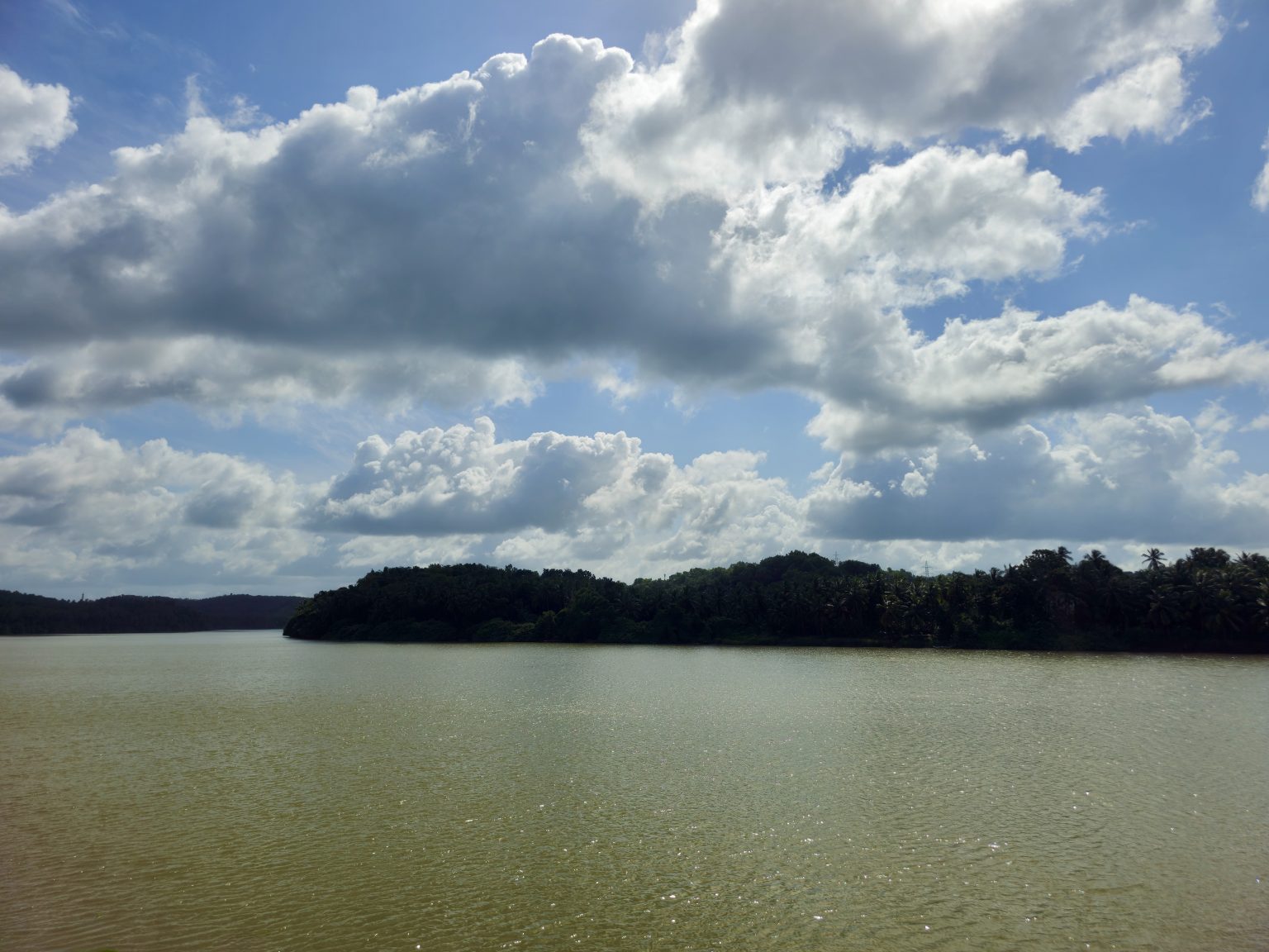 A calm water body stretches toward a green, tree-covered shoreline under a wide sky filled with dramatic clouds. The peaceful landscape, reflections, and cloud formations create a scenic outdoor photograph. Location: Manakkadavu, Kozhikode, Kerala.