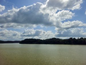 A calm water body stretches toward a green, tree-covered shoreline under a wide sky filled with dramatic clouds. The peaceful landscape, reflections, and cloud formations create a scenic outdoor photograph. Location: Manakkadavu, Kozhikode, Kerala.