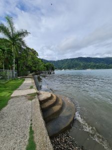 A quiet waterfront walkway curves alongside a tropical bay, where gentle waves, lush vegetation, and anchored sailboats create a peaceful coastal scene.
