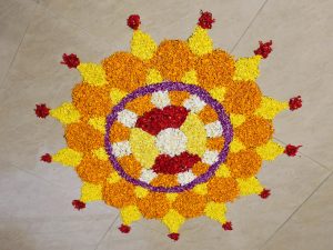 A colorful circular Pookkalam arranged on a light tiled floor using orange, yellow, purple, white, and red flower petals. The layered floral pattern forms a decorative traditional design for an Onam celebration. Location: Perumanna, Kozhikode, Kerala.