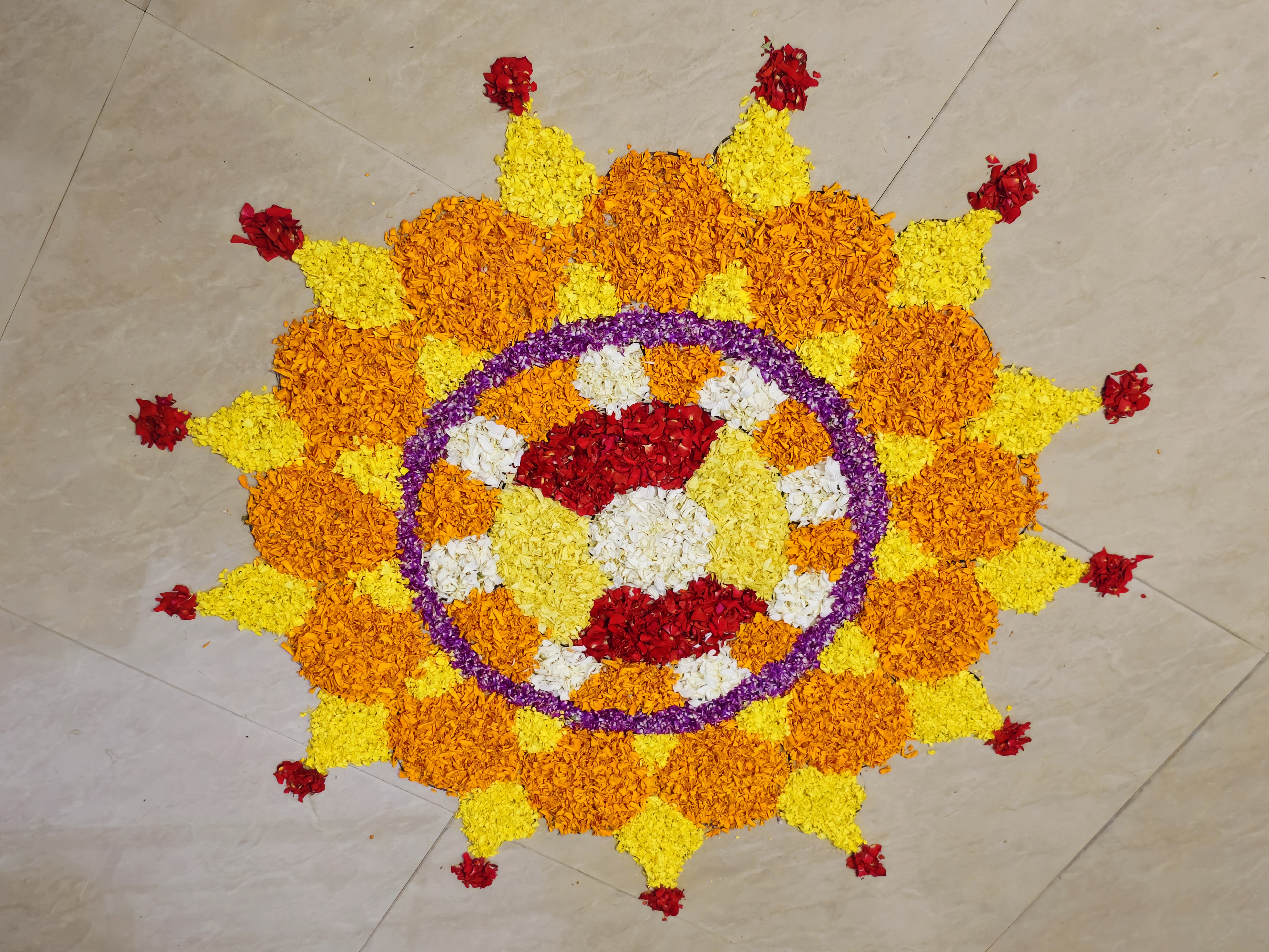 A colorful circular Pookkalam arranged on a light tiled floor using orange, yellow, purple, white, and red flower petals. The layered floral pattern forms a decorative traditional design for an Onam celebration. Location: Perumanna, Kozhikode, Kerala.