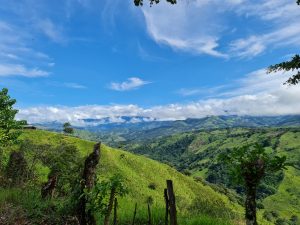 Rolling green hills stretch toward a distant mountain range, where low clouds drift across the landscape beneath a vivid blue sky
