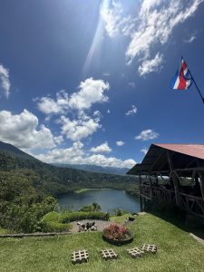 A peaceful lake surrounded by green mountains under a blue sky with white clouds. A wooden shelter, garden, and flag are in the foreground.