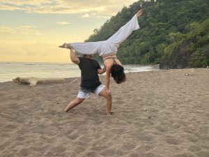 A man is lifting a woman into the air in a playful pose on a sandy beach. The woman is upside down, her legs extended upwards, while the man supports her waist, maintaining balance. The scene is set during sunset, with a colorful sky painted in soft hues of blue and orange. 
